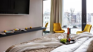 Ein Hotelzimmer mit Bett, Fernseher, Tisch mit Flasche und Gläsern, gelben Stühlen und Blick auf Gebäude.
