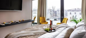 A hotel room with a bed, a TV, a table with a bottle and glasses, yellow chairs, and a view of buildings. - Rajska 3 by Fragola Apartments (Kraków)