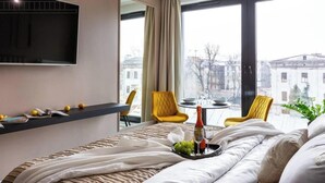 Ein Hotelzimmer mit einem Bett, einem kleinen Tisch mit Flasche und Gläsern, einem Sessel, einem an der Wand montierten Fernseher und einem Fenster mit Blick auf die Stadt.