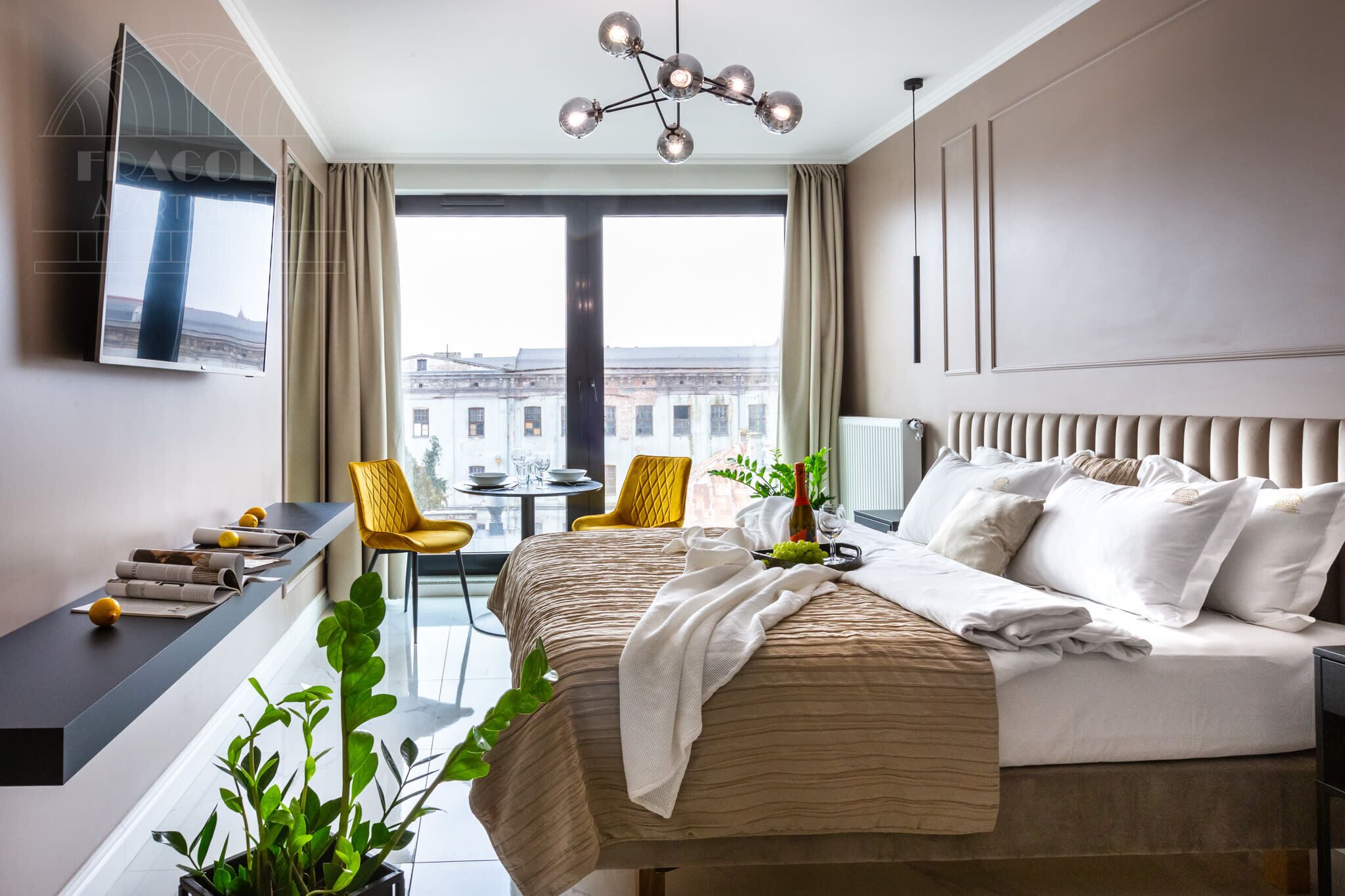 A modern hotel room with a large bed, a TV mounted on the wall, a dining area with a table and chairs, and a view of the cityscape.