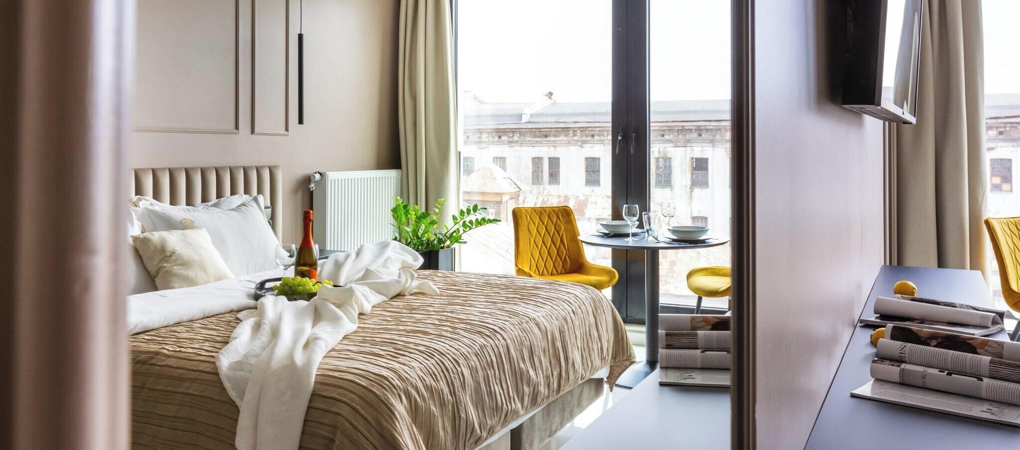 Habitación de hotel con una cama, una mesita con una planta, una silla amarilla y vistas a edificios.
