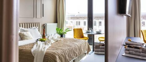 Habitación de hotel con una cama, una mesita con una planta, una silla amarilla y vistas a edificios.