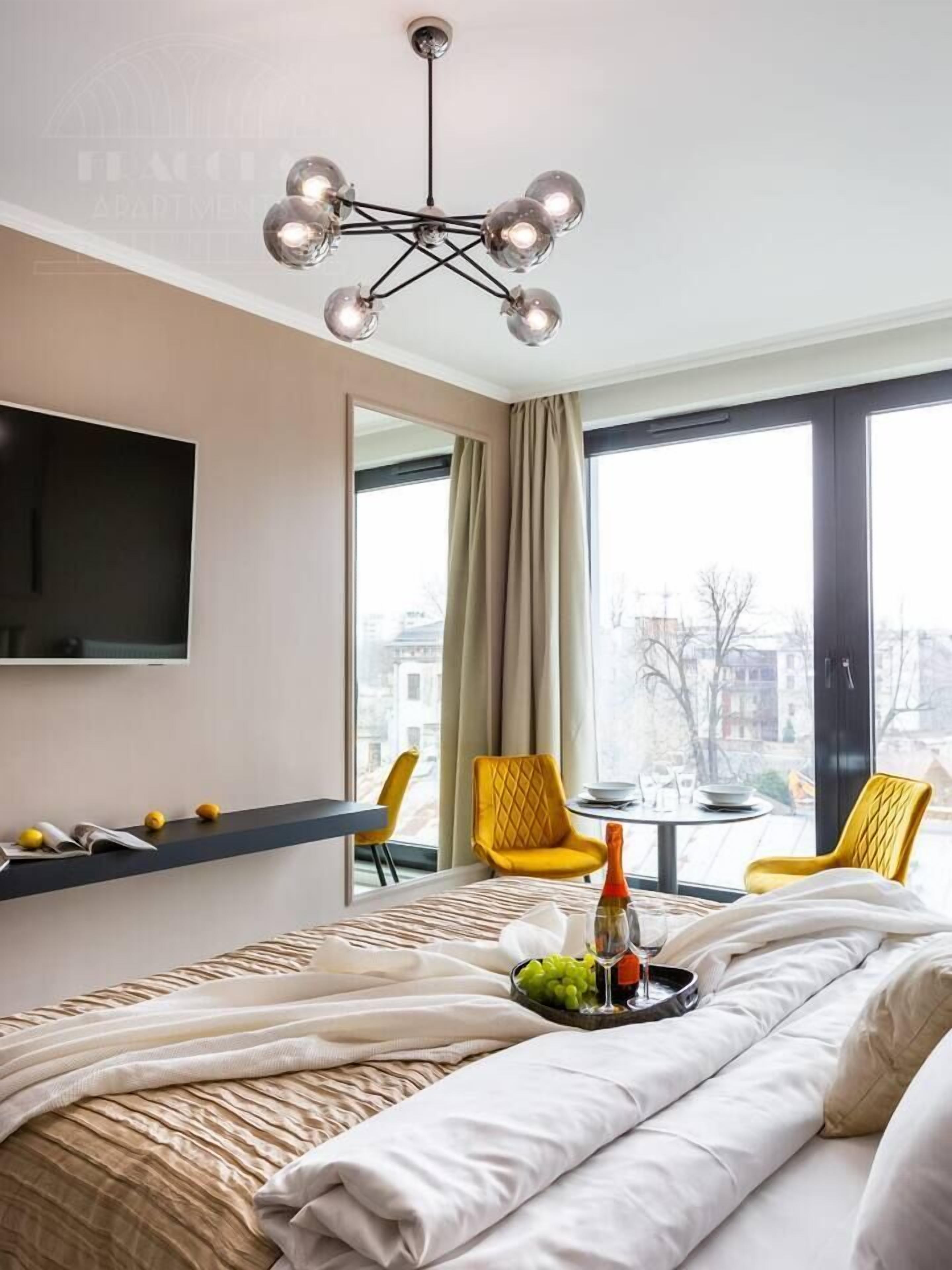 Una habitación de hotel moderna con cama, televisor, candelabro, ventana con vistas y una pequeña mesa con sillas.