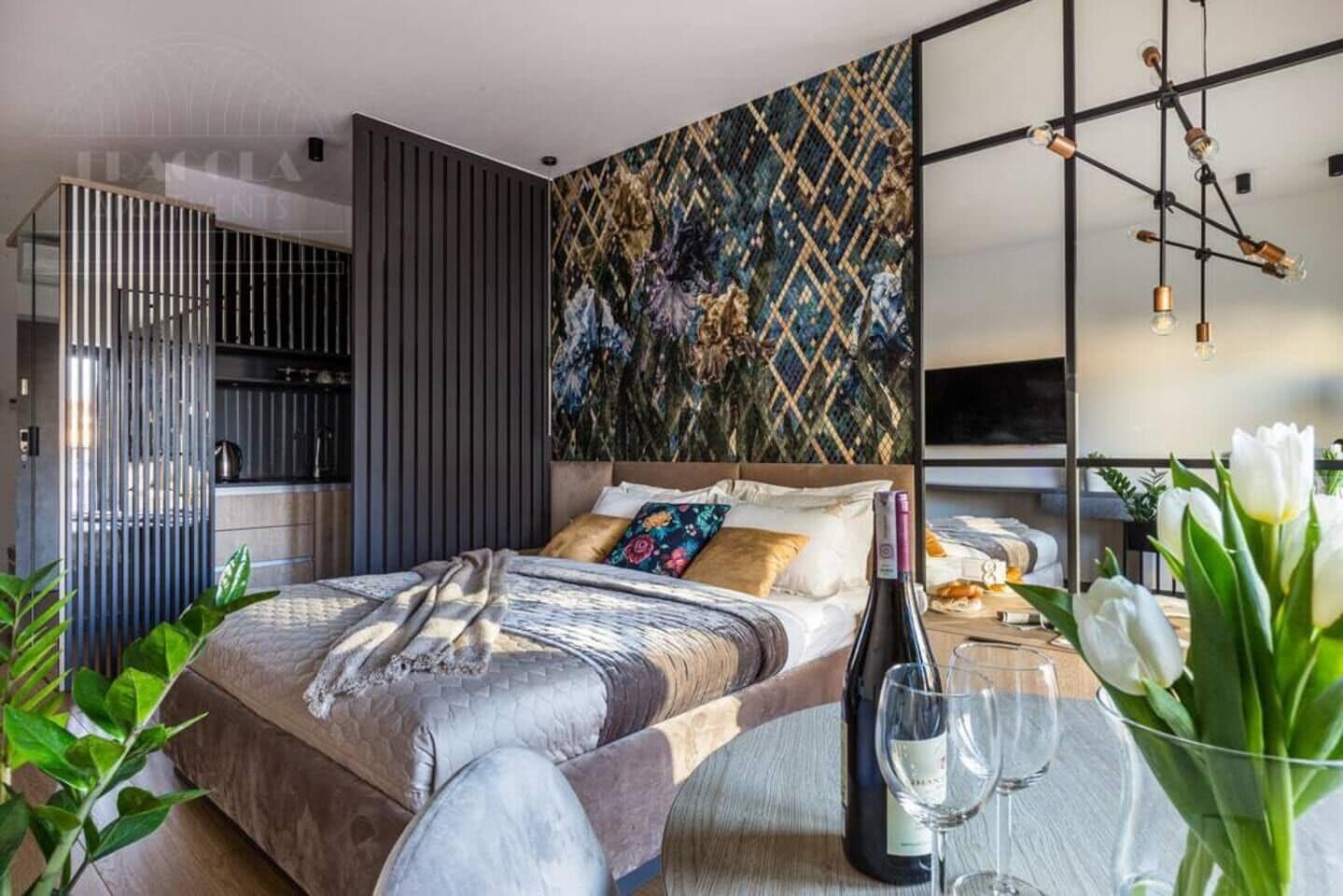 A modern bedroom with a bed, a dining table with a bottle and glass, a kitchen area, and a decorative wall.