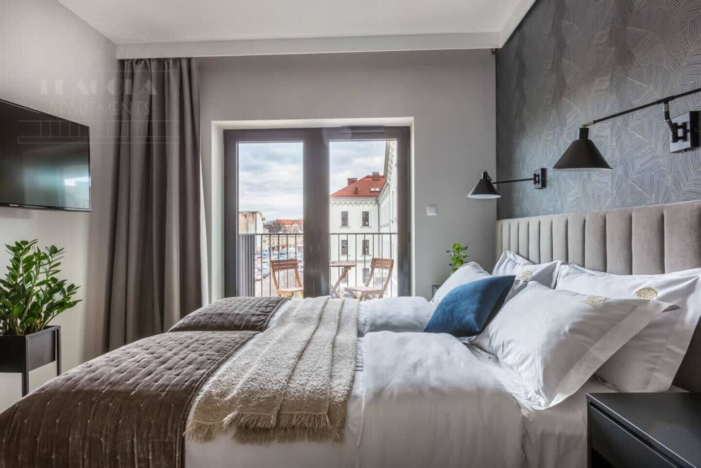 A modern bedroom with a large bed, a TV mounted on the wall, and a balcony with a view of buildings.