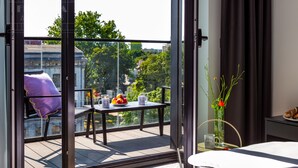 Ein modernes Hotelzimmer mit Balkon, einem Esstisch mit Obst und Kaffeetassen, einer Vase mit Blumen sowie einem Bett mit weißem Kopfteil.