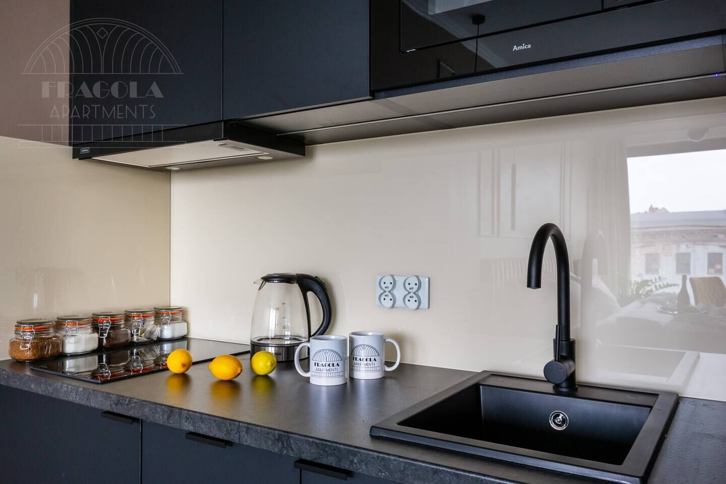A modern kitchen with a black sink, a kettle, and a coffee maker. There are jars of spices and lemons on the counter.