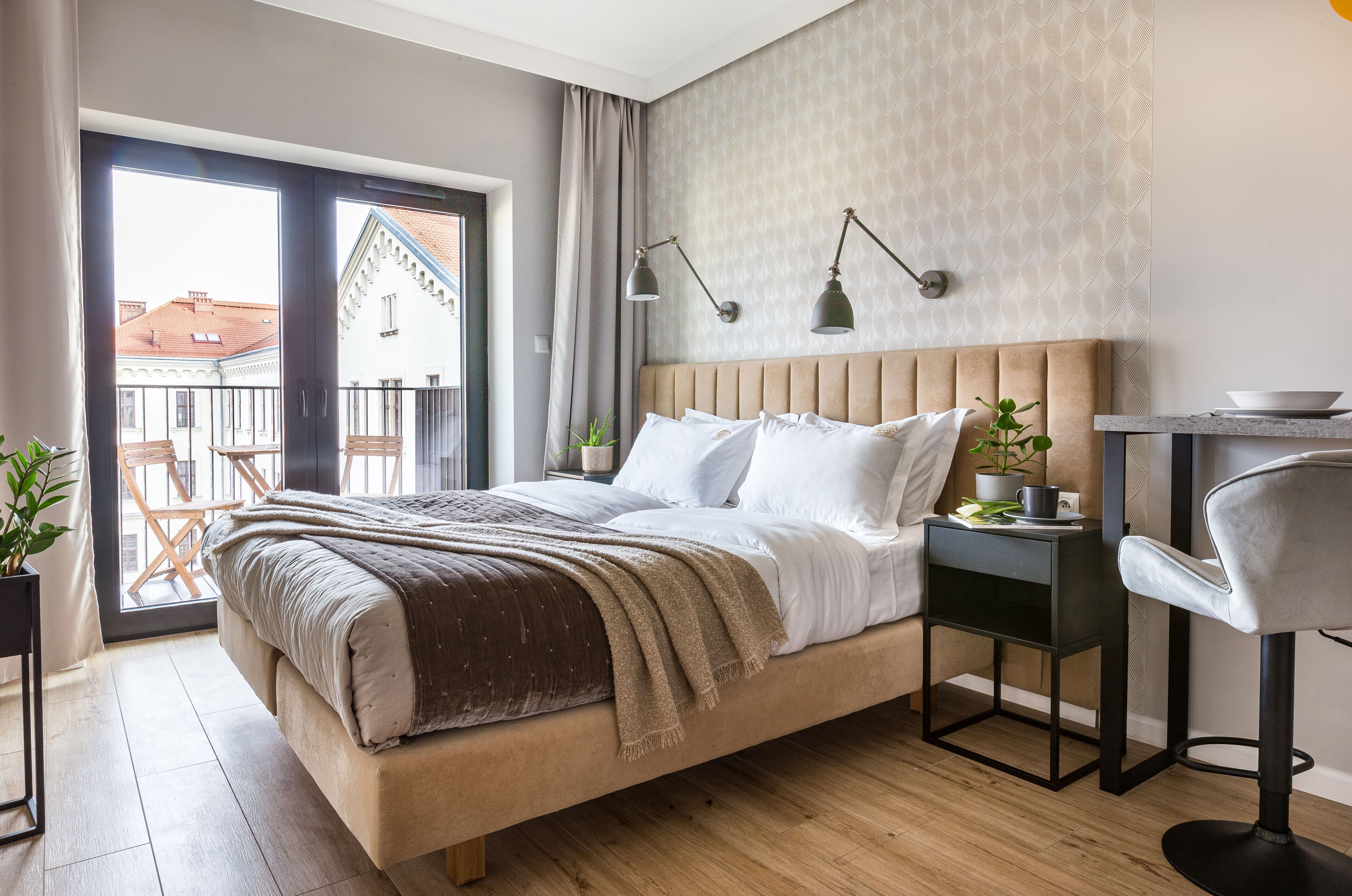 A bedroom with a large bed, a small table with a bowl, a chair, and a balcony with a view of buildings.