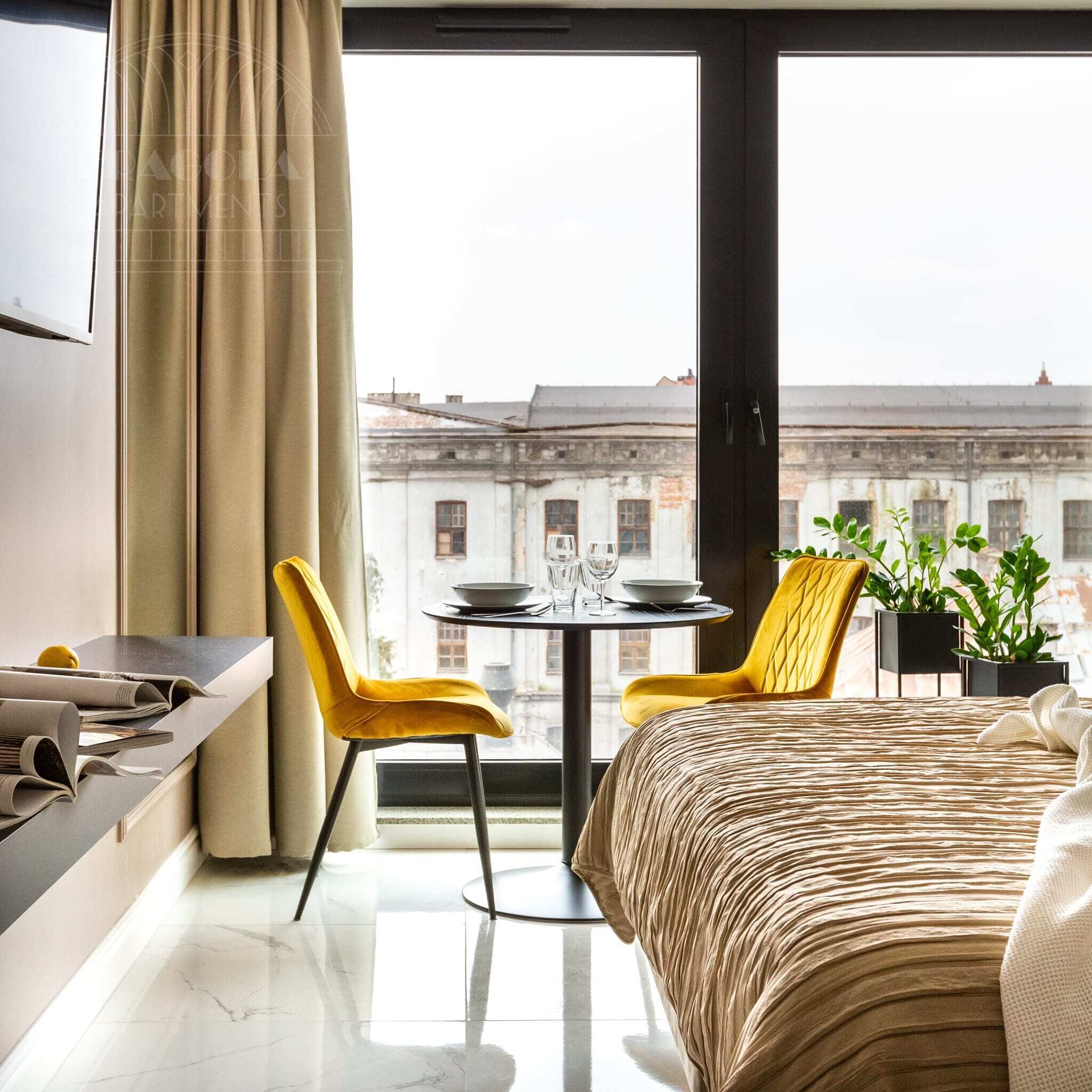 A hotel room with a bed, a table with chairs, a TV, and a view of buildings.