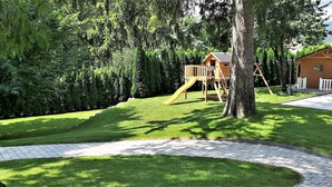 A playground with a wooden slide and climbing structure, surrounded by lush greenery and a paved walkway. - Vila Ključe Apartments Bled (Bled)