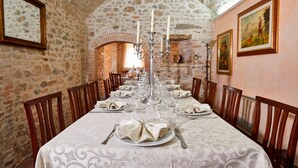 Une longue table à manger, recouverte de nappes blanches, agrémentée de bougies et de assiettes, dans une pièce aux murs de pierre et aux chaises en bois.
