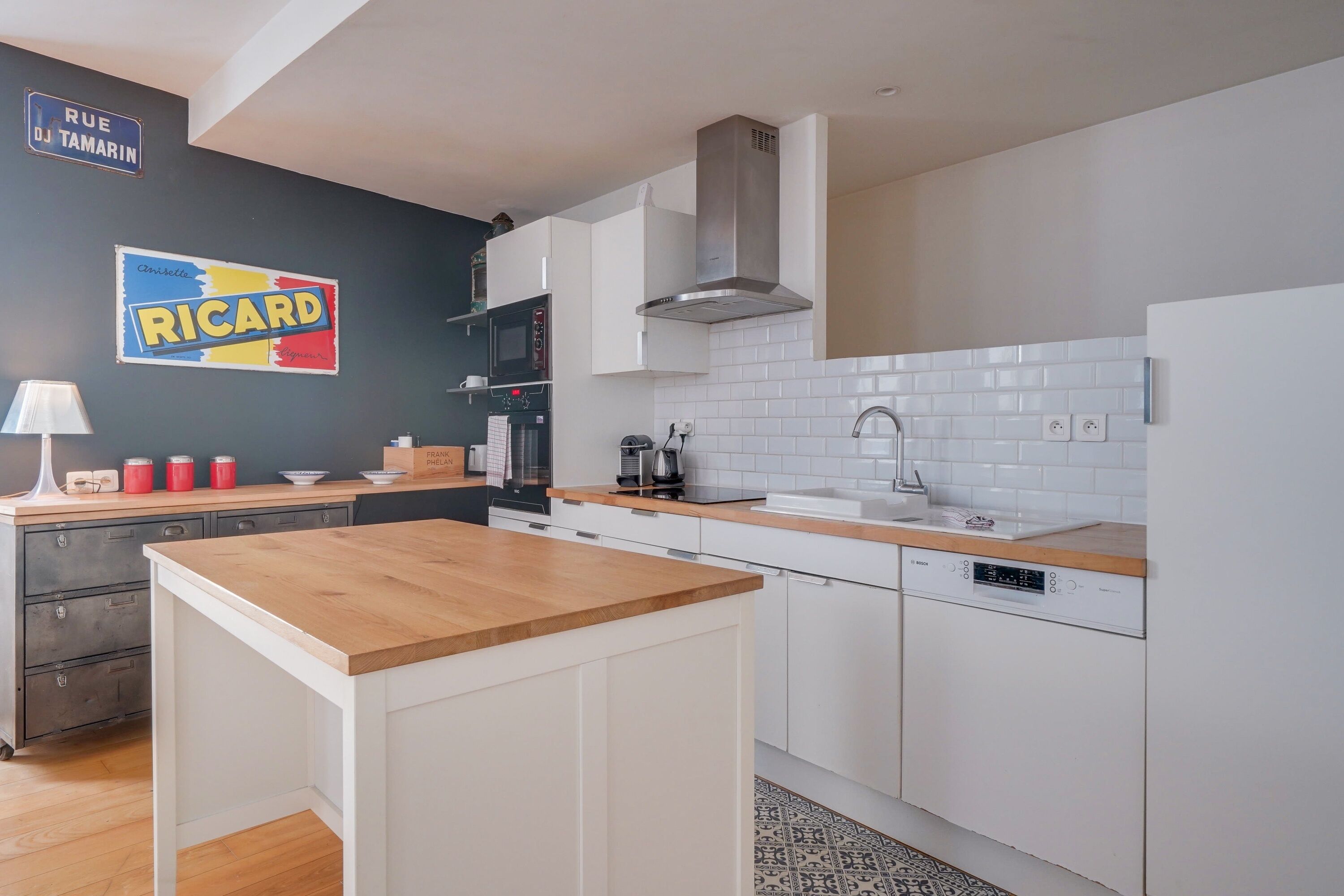 Een moderne keuken met witte kasten, een houten aanrecht, een Bosch vaatwasser, en een zwarte muur met een 'RICARD' bord.