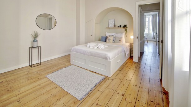 Ein Schlafzimmer mit einem weißen Bett, einem runden Spiegel, einem kleinen Beistelltisch mit einer Pflanze und einem Flur, der in einen anderen Raum führt.