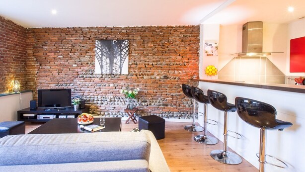 A modern living space with a brick wall, a flat-screen TV, a dining area with bar stools, and a kitchen with a stainless steel range hood. - LE ROSA BONHEUR - One Bedroom Apartment, Sleeps 4 (Toulouse)
