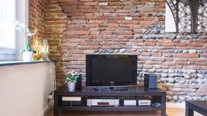 A living room with a brick wall, a flat-screen TV on a wooden stand, a potted plant, and a decorative wall piece. - LE ROSA BONHEUR - One Bedroom Apartment, Sleeps 4 (Toulouse)