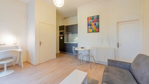 A modern living room with a grey sofa, a white coffee table, and a dining area with a small table and chairs. There is a kitchen in the background with dark cabinets and a colorful wall art piece.