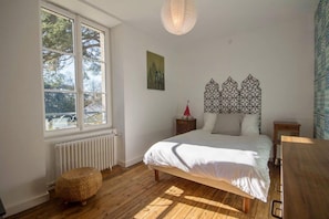 Um quarto com uma janela grande, um radiador, uma cama com cabeceira decorativa, uma pequena mesa de cabeceira e um banquinho de fibra natural.