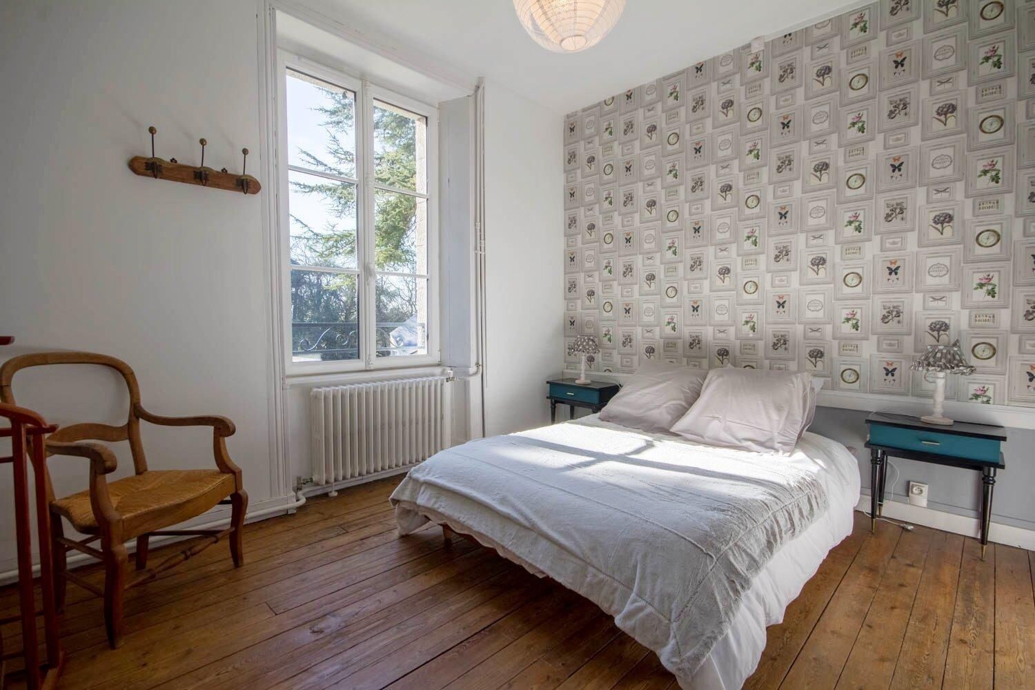 A bedroom with a wooden floor, a bed with white bedding, a chair, a nightstand with a lamp, and a window with a view of trees.