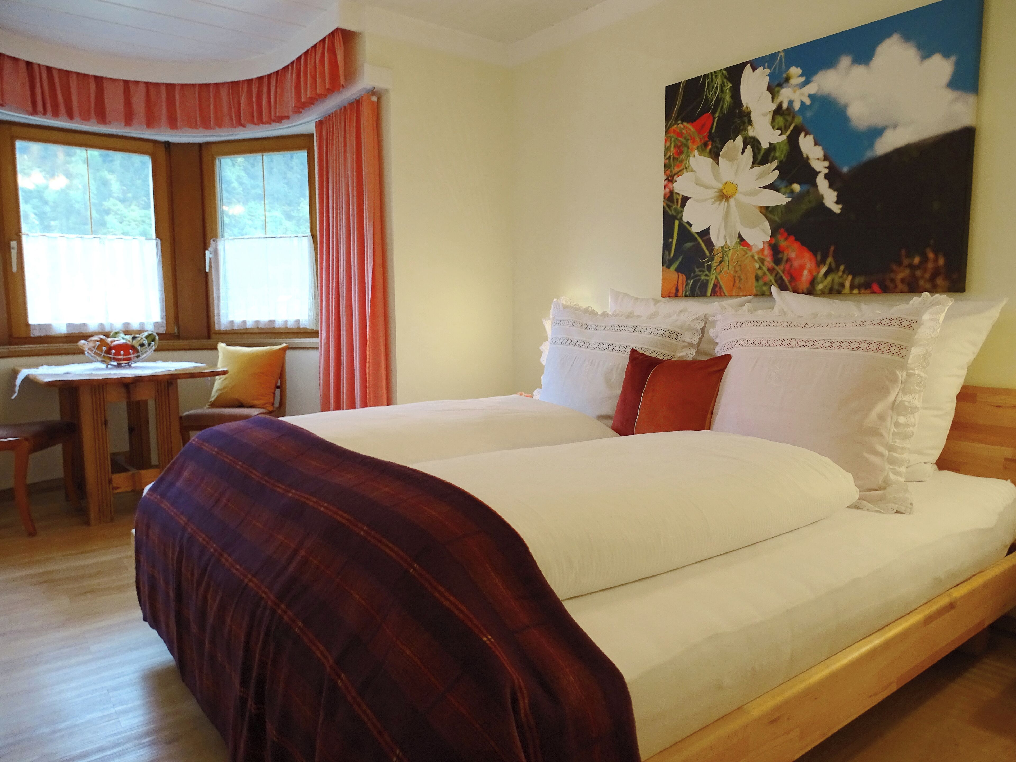 A bedroom with a large bed, a wooden headboard, a painting of flowers, and a window with curtains.