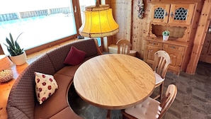 L’intérieur chaleureux d’un chalet avec une table ronde en bois, un banc intégré agrémenté de coussins, des armoires en bois et une grande fenêtre offrant une vue imprenable.