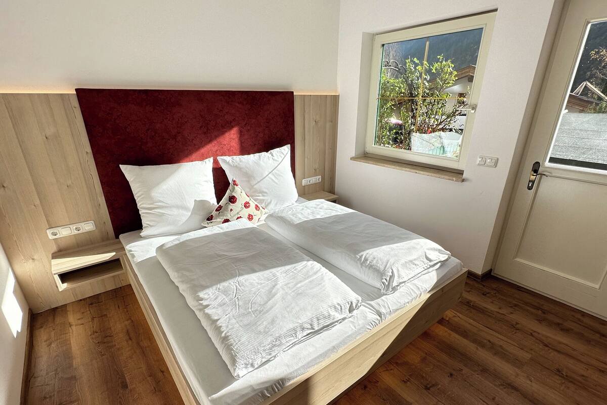 Un lit avec du linge de lit blanc, une tête de lit rouge et une fenêtre donnant sur un paysage verdoyant.