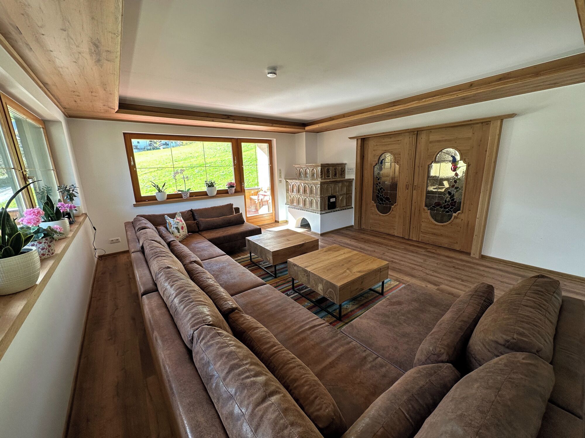 A spacious living room with a large brown sectional sofa, a wooden coffee table, and a view of a green garden through large windows.