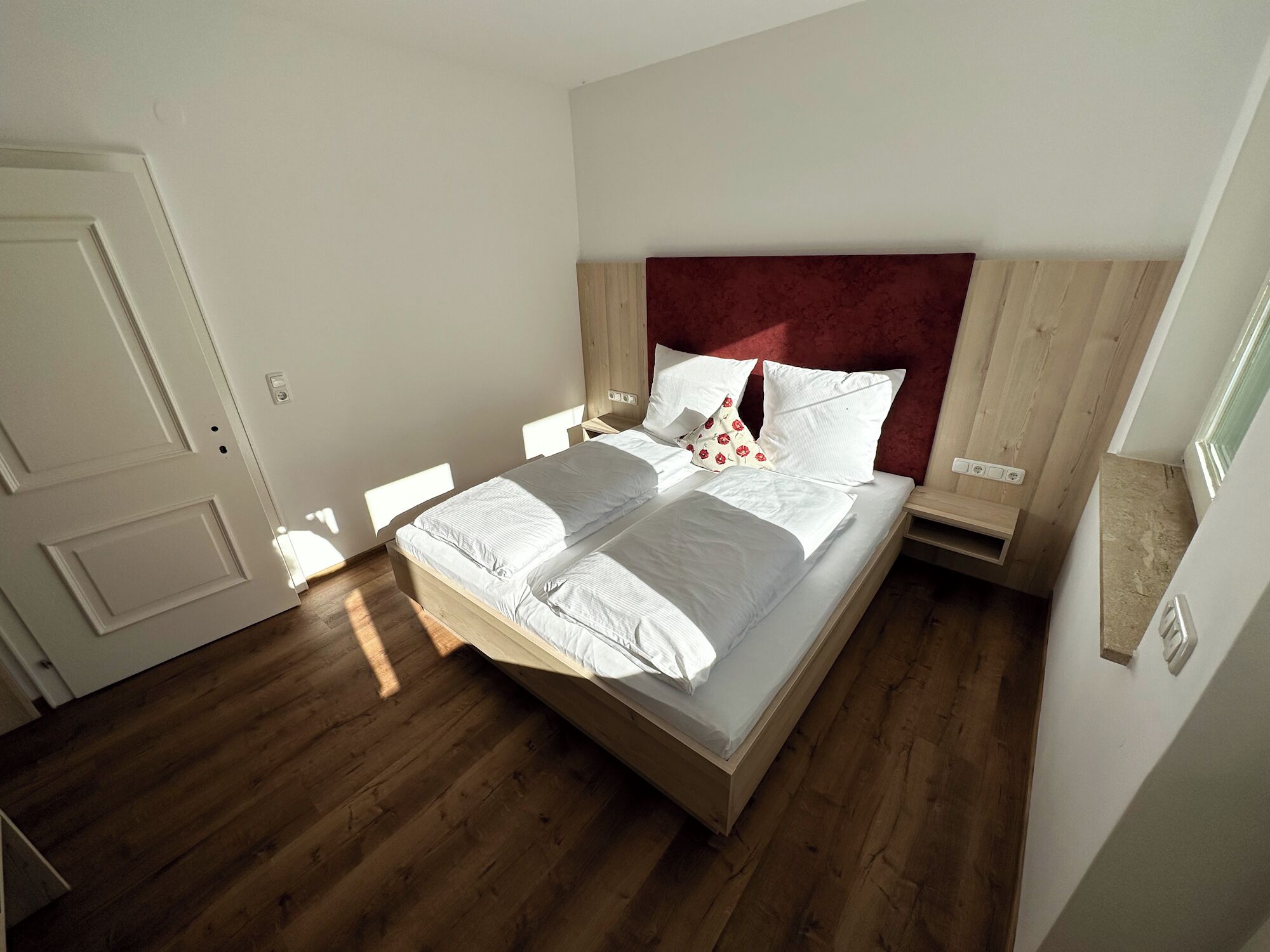 Un lit bien fait, avec du linge de lit blanc et une tête de lit rouge, dans une pièce au plancher en bois et aux murs blancs.