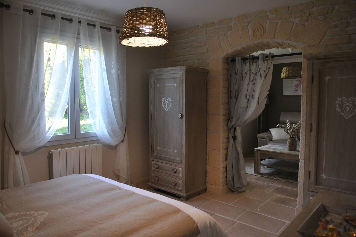 A bedroom with a bed, a wardrobe, a window with sheer curtains, and a stone wall.
