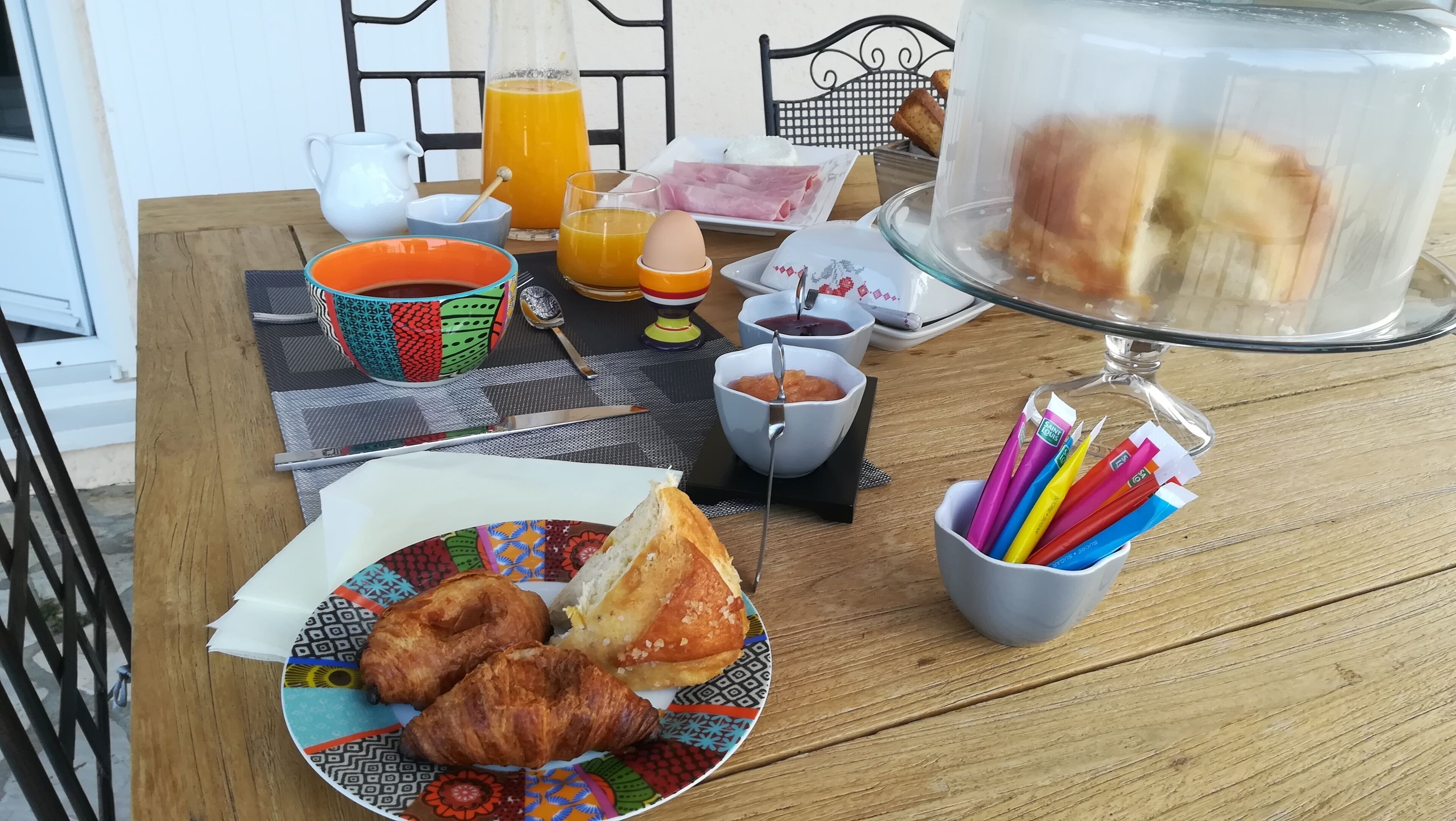 A breakfast table with croissants, a plate of bread, a glass of orange juice, a pitcher, a bowl of jam, and a cup of coffee.