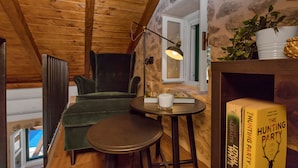 A cozy room with wooden ceiling beams, a green armchair, a small round table, a floor lamp, and a bookshelf with books.