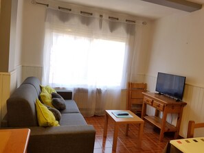 A living room with a sofa, a television on a wooden stand, a small table, and a window with curtains. - 4/5 CONSUEGRA 1 -apt 1 hab- 5 pers. 150 m piste- Pas de la Casa 005544 (PAS DE LA CASA)
