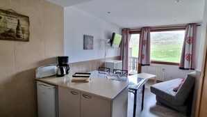 Eine kleine Küche mit Kaffeemaschine, Toaster und einem kleinen Kühlschrank. Es gibt einen Essbereich mit Tisch und Stühlen sowie ein Fenster mit Vorhängen.