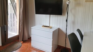 A room with a flat-screen TV mounted on a white wall, a white chest of drawers, a standing lamp, and a view of a wooden deck through a glass door. - GRIZZLY 203-apt-1 hab 5 pers-balco-10 m piste-Pas de laCasa-005865 (Pas de la Casa)