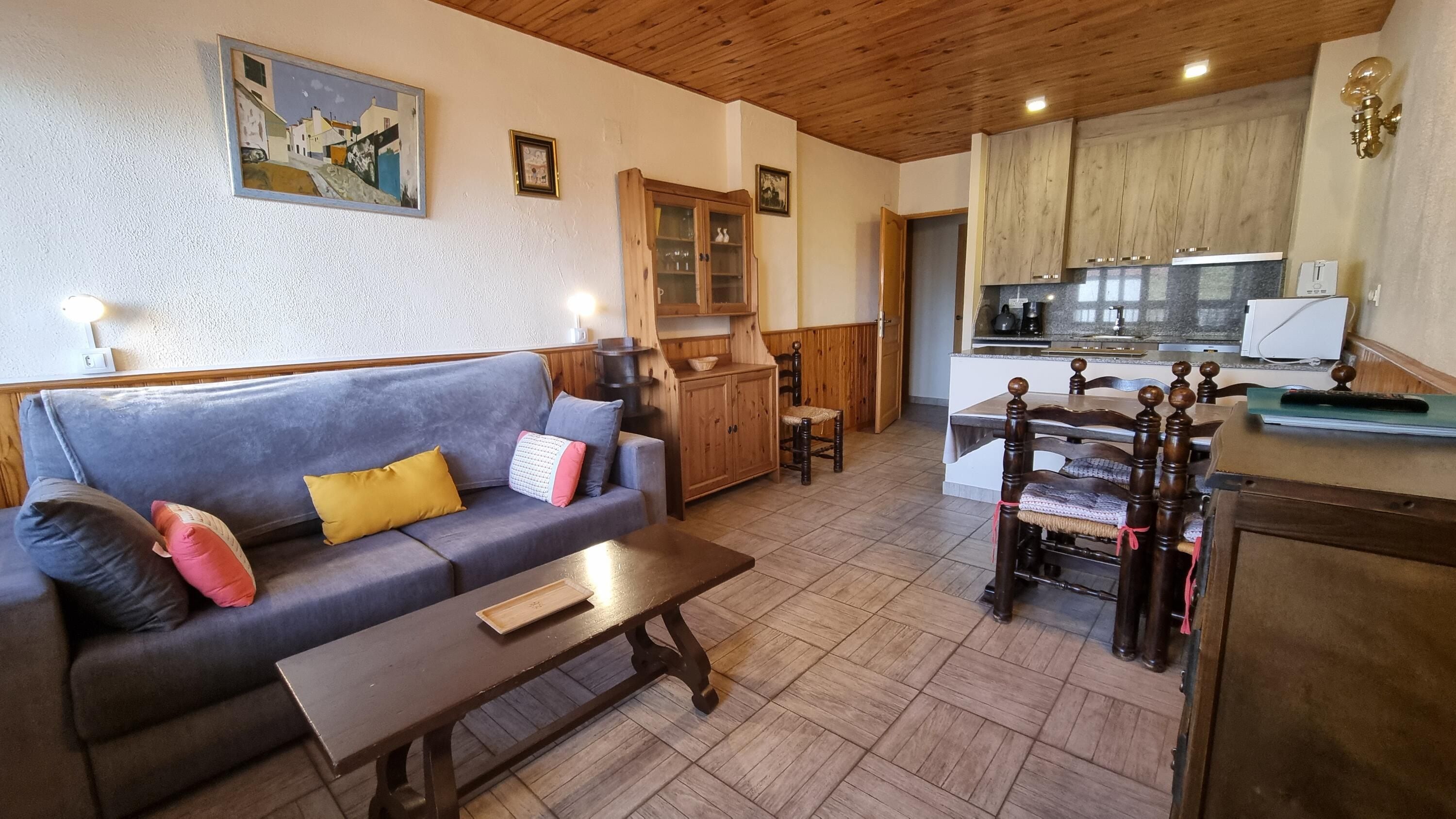 A living room with a sofa, a wooden table, and a kitchen area with cabinets and a countertop.