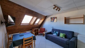 Ein gemütliches Zimmer mit schrägverdeckter Decke, Holzwänden, einem Esstisch mit Stühlen, einer Couch mit Kissen und einem an der Wand befestigten Fernseher.
