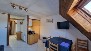A compact room with a dining table, chairs, a TV mounted on the wall, a kitchenette, and a window with a wooden beam. - CANIGOU 67-estudi- 4 pers.-piste- Pas de la Casa -006053 (PAS DE LA CASA)
