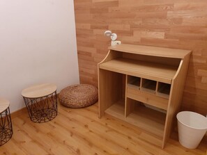 A small wooden room with a wooden floor, a white wall, a wooden bench, a small round table, a woven stool, and a white trash bin.