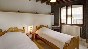 Een kamer met twee houten bedden, een plank met boeken, een raam met gordijnen en een radiator.