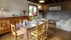 Een eethoek met een houten tafel voor vier personen, omringd door houten stoelen. Op de tafel staat een vaas met bloemen, en links staat een lamp. De kamer heeft een houten plafond en een bank op de achtergrond.