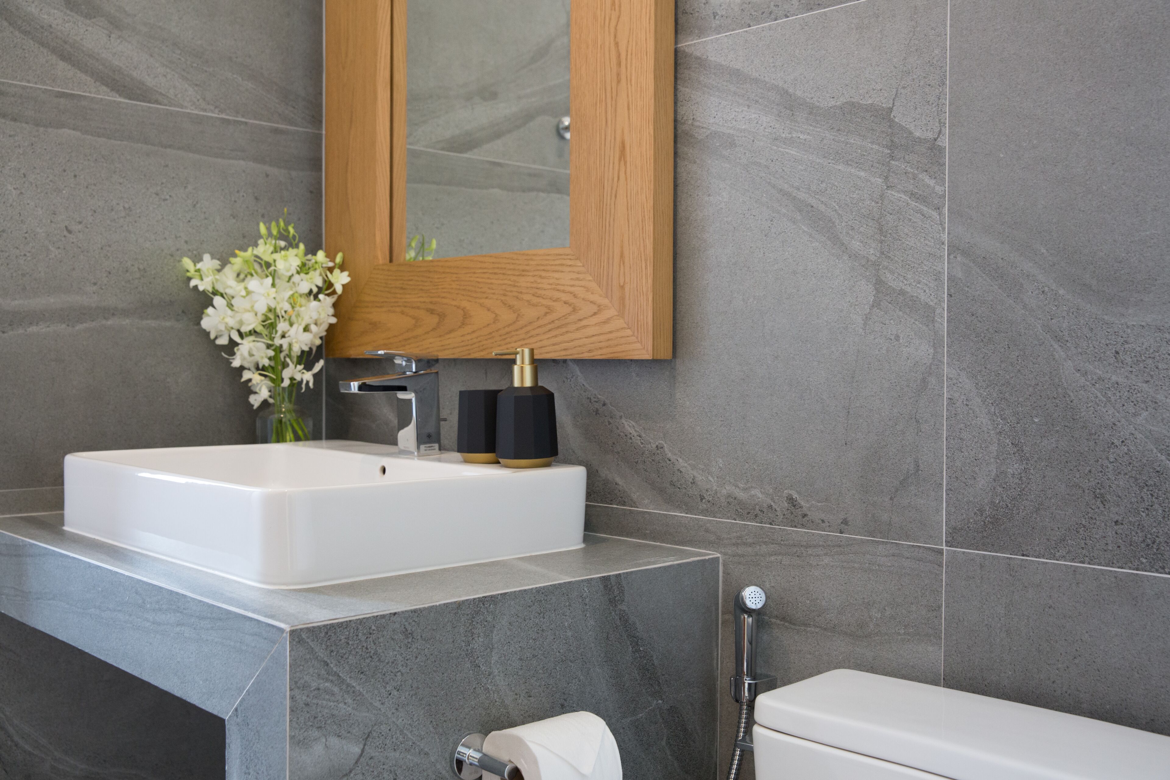 A modern bathroom with a large white sink, a wooden-framed mirror, and grey stone tiles.