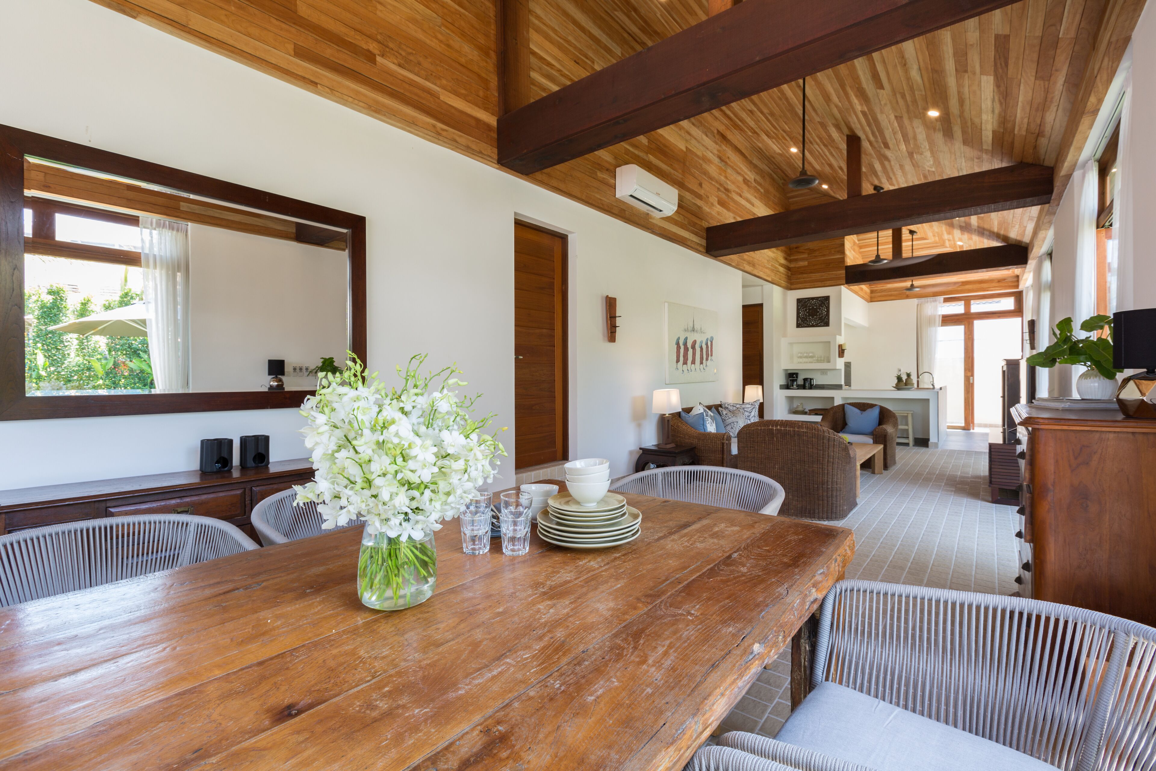 A spacious dining area with a wooden table, white flowers, and a view of the kitchen in the background.