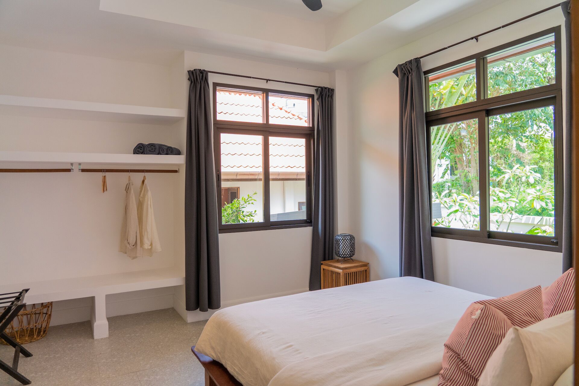 A bedroom with a bed, a bench, a small table, and two windows with curtains.
