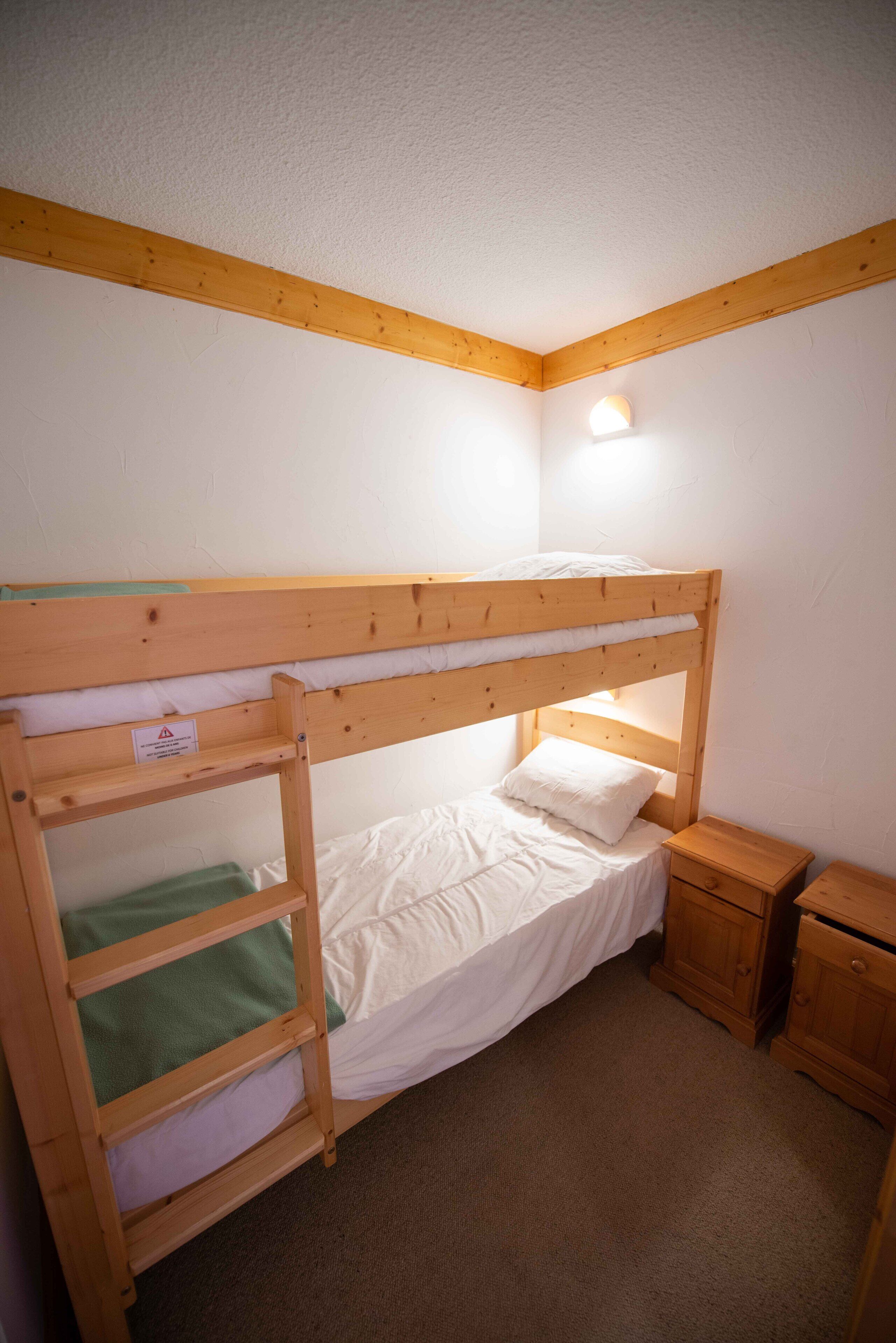 A bunk bed with a green shelf, a bedside table, and a nightstand in a corner room with white walls and wooden beams.