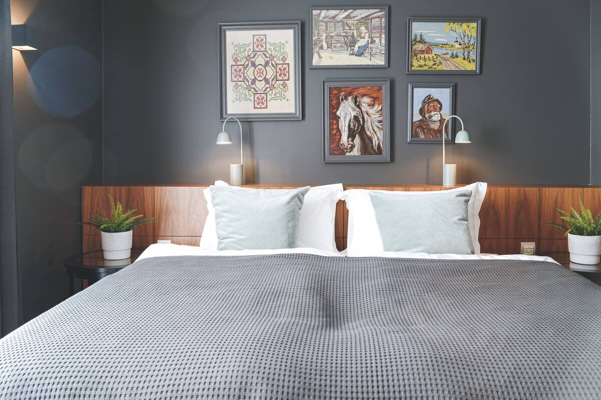 Quarto moderno com cabeceira de madeira, colcha estampada em cinza, duas mesas de cabeceira com luminárias e quatro obras de arte emolduradas na parede.