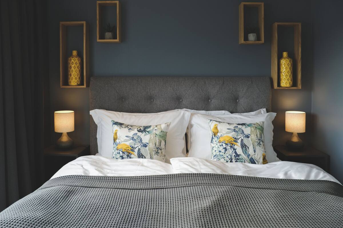 Cama bem arrumada com roupa de cama branca e almofadas decorativas, com duas luminárias de cabeceira sobre mesas de cabeceira de madeira.