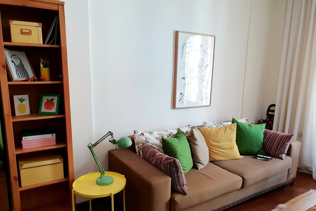 A living room with a brown sofa, a small yellow table, and a bookshelf with various items.