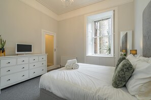 A bedroom with a large bed, a dresser with drawers, a television on top, a window with a view of buildings, and a radiator.