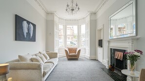 Een ruime woonkamer met een beige bank, een fauteuil, een open haard en een groot raam met uitzicht op de straat.