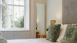 Een opgemaakt bed met een witte dekbedovertrek, een groene gebogen kussen en een passend kussen op een houten nachtkastje. Een spiegel hangt aan de muur naast het bed.