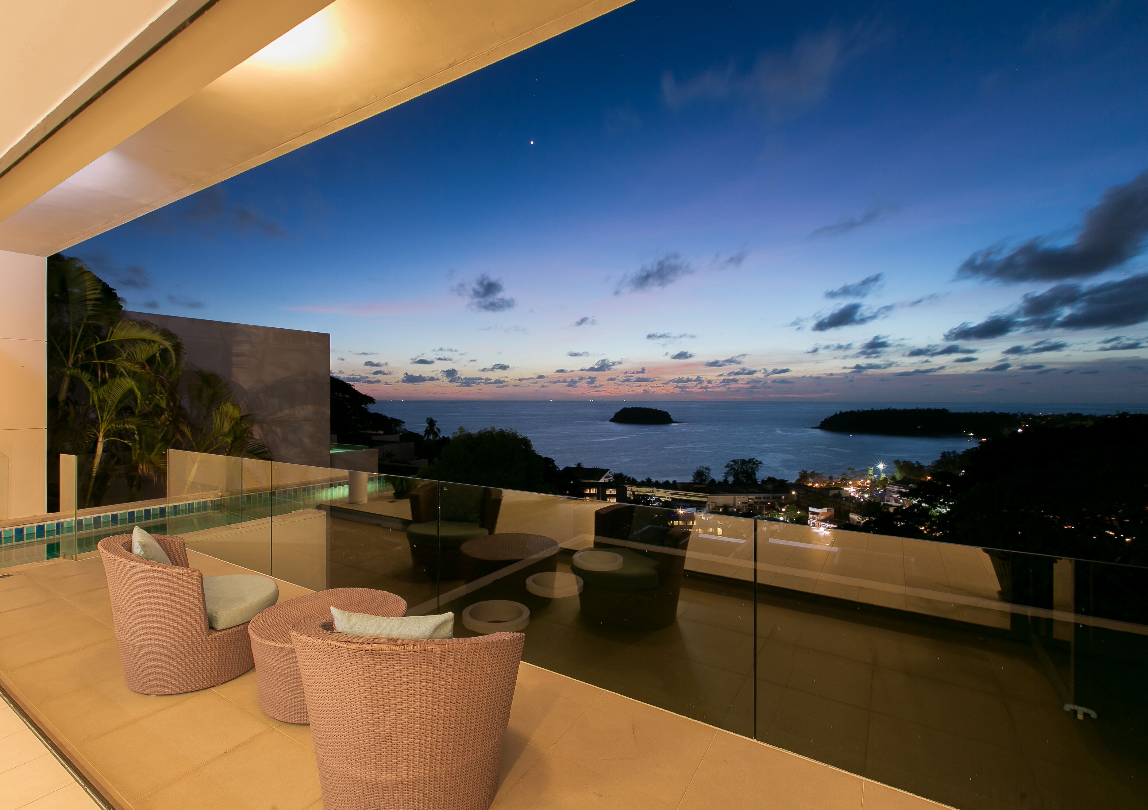 A balcony with wicker furniture, a glass railing, and a view of the sea at dusk.